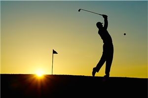 Silhouette of a man hitting a golf ball towards the hole at sunset.